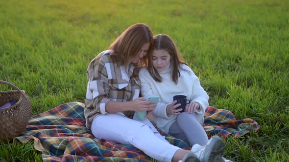 Young mum with a cute daughter use smartphone sit on plaid on a field at sunset