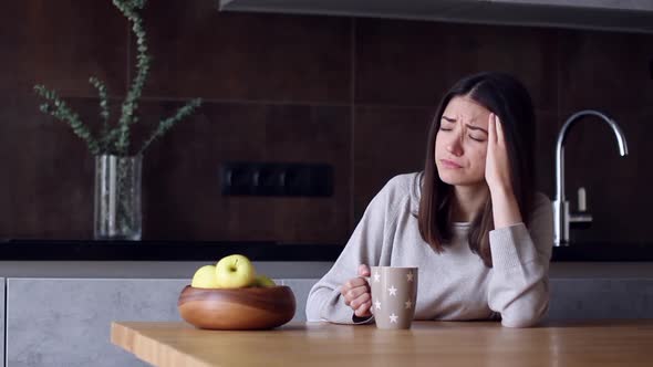 Woman With Headache Sitting In Kitchen