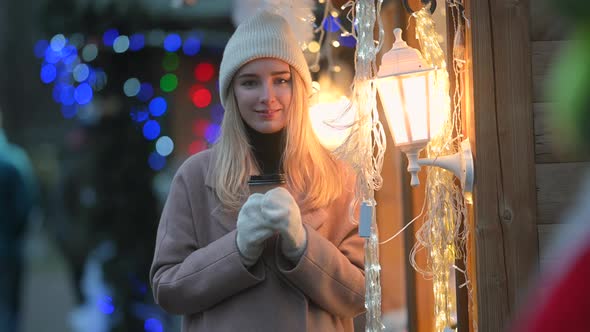 young girl with a cup of hot coffee in her hands stands against the background of Christmas lights
