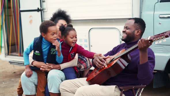 Smiling African father playing guitar to his family near trailer