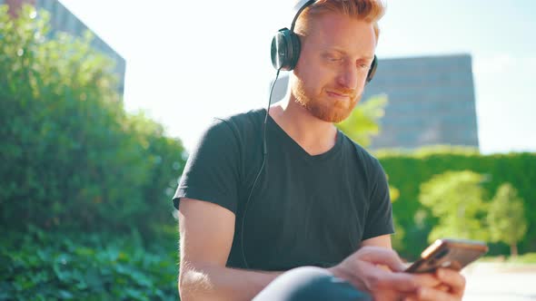 Positive red haired man listening music in headphones and typing by phone