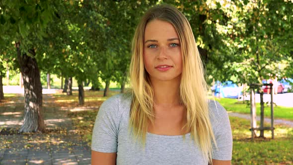 Young Pretty Blond Woman Looks To Camera with Serious Face - Park with Trees in Background