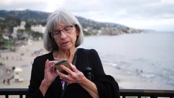 A middle aged woman with white hair and eye glasses texting on her phone on vacation in Laguna Beach