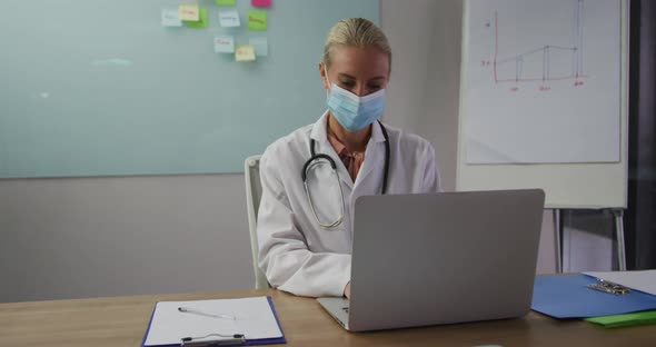 Caucasian female doctor wearing mask sitting at desk in meeting room using laptop