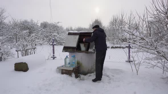 A Man Takes a Bucket of Water From a Well and Pours It Into Plastic Bottles During a Snowfall