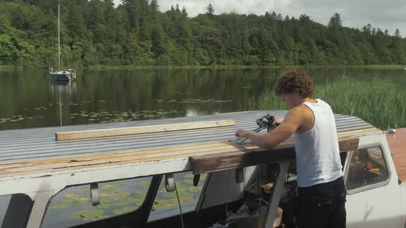 Young man res masking tape after sealing wooden boat roof starboard side.