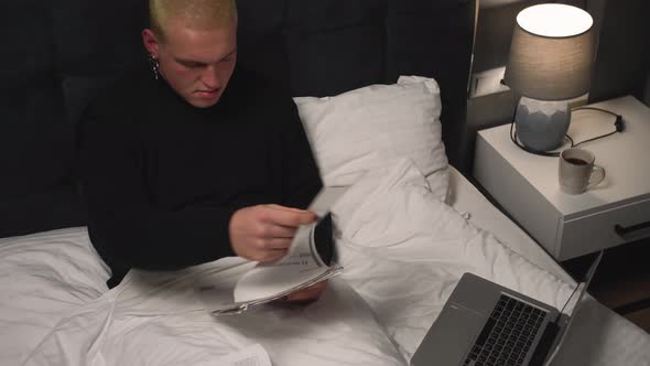 Handsome Homosexual Man Working with Documents Using Laptop on Bed Before Going To Sleep at Home