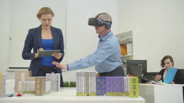 Woman Stands Next To Model of Houses with Tablet in Hands Talks with Colleague in Virtual Glasses in