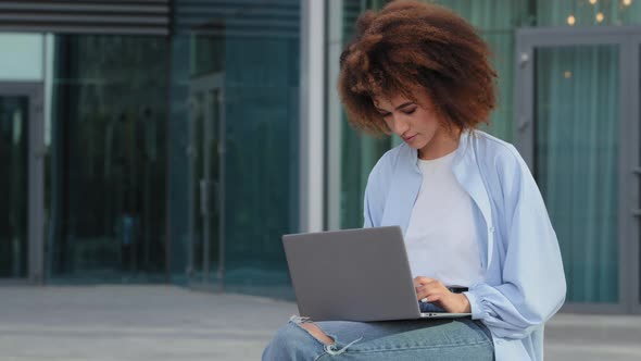 Young Curly African American Girl Woman with Afro Hairstyle Sitting Outdoors on Street in City in