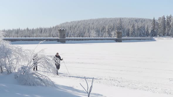 Two Crosscountry Skiers Walk Along the Shore of a Frozen Lake  a Dam and a Forest in Background