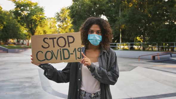 Darkskinned Woman in Protective Mask Showing Cardboard Poster with Inscription Stop Covid19 Posing