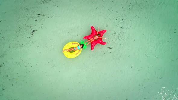 Aerial view of two girls floating on inflatable mattress in transparent sea, holding each other.