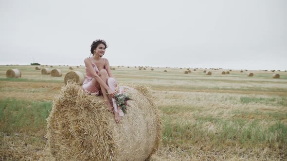 Lady in Elegant Dress and Accessories Poses with Joy on Haystack in Windy Field