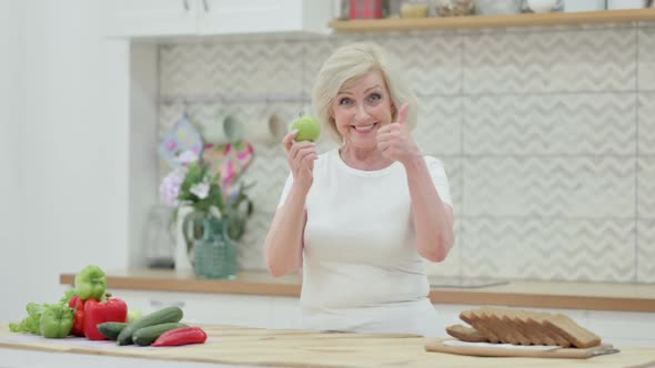 Old Woman Showing Thumbs Up While Holding Apple in Kitchen