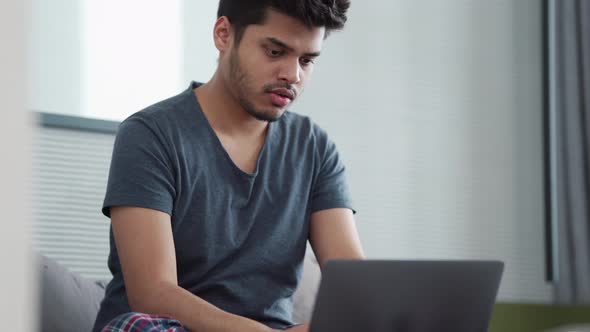Handsome Indian man wearing pajama typing by laptop and looking to the side