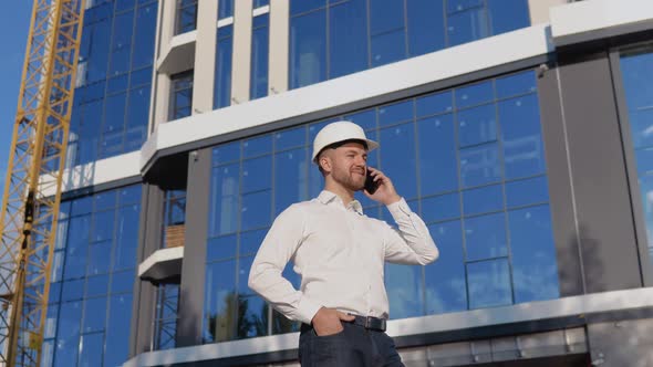 Engineer Architect in a White Shirt and Helmet on the Background of a Modern Glass Building Speaks