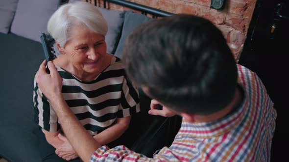 Happy Pensioner Mother Sitting on a Sofa and Having Her Hair Combed By Her Middleaged Son