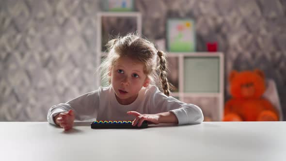 Smart Little Girl with Toy Abacus Does Sums Sitting at Table