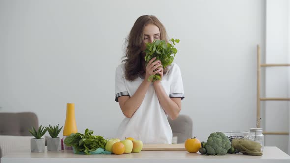 Young Smiling Woman Sniffing Fresh Greens Standing at the Table in Modern Kitchen