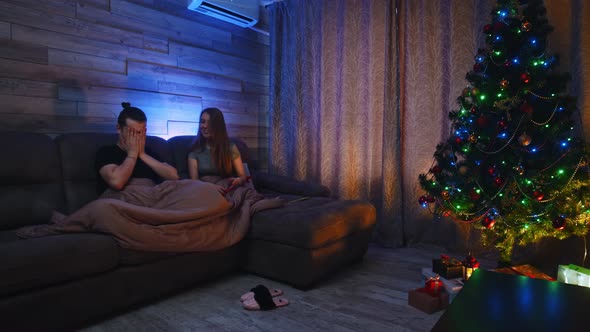 Young Couple Sitting on a Couch Under a Blanket  the Woman Gives Her Boyfriend a Christmas Gift