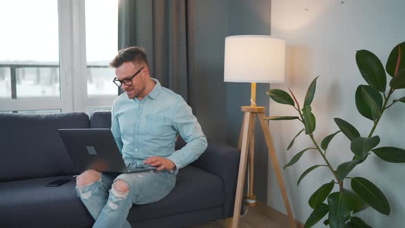 Casual Dressed Man with Glasses Sitting on the Couch and Using Laptop to Make a Video Call From Home