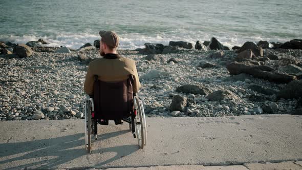 Alone Male Wheelchair User Is Sitting Calmly on Seacoast and Relaxing Back View