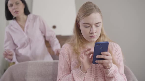 Camera Approaching To Worried Caucasian Girl Typing on Smartphone As Her Mother Looking at Screen