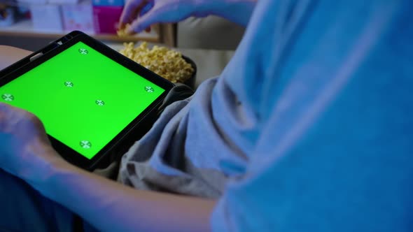 Close up, A man holding tablet with green touch screen and eating popcorn on sofa at home.