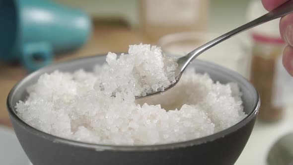 Bowl full of coarse sea salt. A spoon scoops up crystals in slow motion on a table