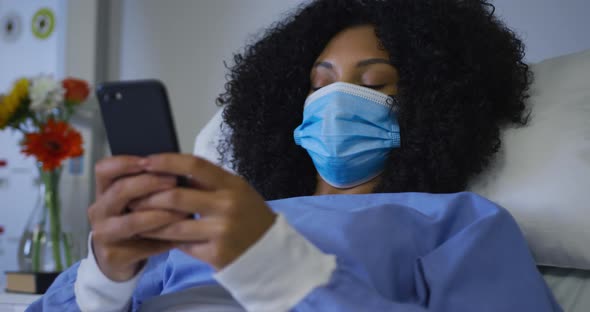 African american female patient wearing face mask lying on hospital bed using smartphone