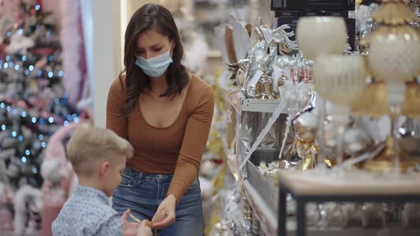 A Woman in a Medical Mask with a Boy in a Shopping Mall Choose Home Decorations for Christmas