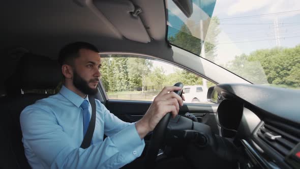 Young Bearded Businessman Driving Car on City Road in Bright Summer Day.