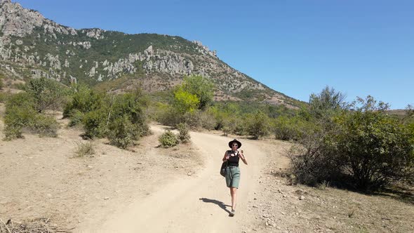 Girl Traveler Walks Alone in a Hat on the Road in the Mountains