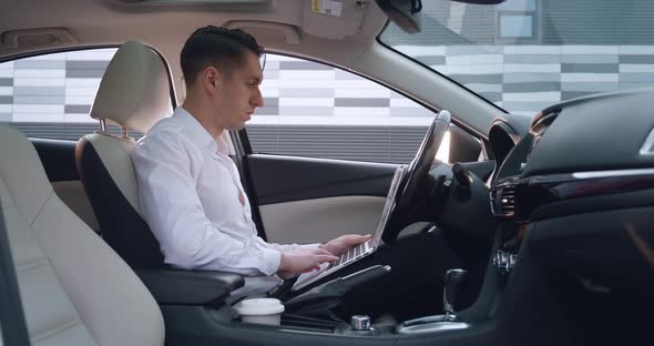 Portrait of Young Male Entrepreneur Sits in the Driver's Seat in a Car and Uses Laptop