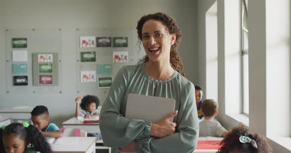 Video of happy caucasian female teacher holding laptop in class of diverse pupils