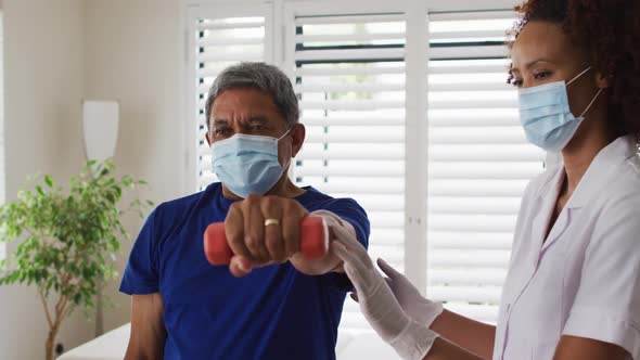 Mixed race female physiotherapist wearing mask helping senior exercise using dumbbells