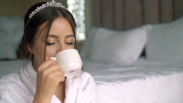 Very beautiful girl with a crown on her head drinking her morning coffee in the bedroom.