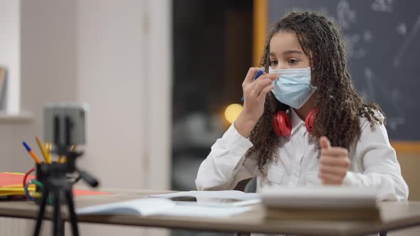 Portrait of Smart Positive African American Schoolgirl in Covid Face Mask Recording Educational