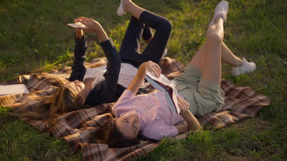 Wide Shot Two Relaxed Teenage Girls Enjoying Hobbies Lying on Sunny Park Lawn Outdoors at Golden