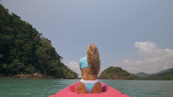 Sexy Young Woman in Blue Swimsuit Swings Feet in Water on Pink Canoe on Sea Against Hills Backside