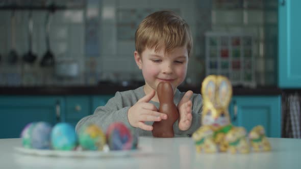 Blonde boy eating Easter chocolate bunnies with dirty face at home on blue kitchen. 