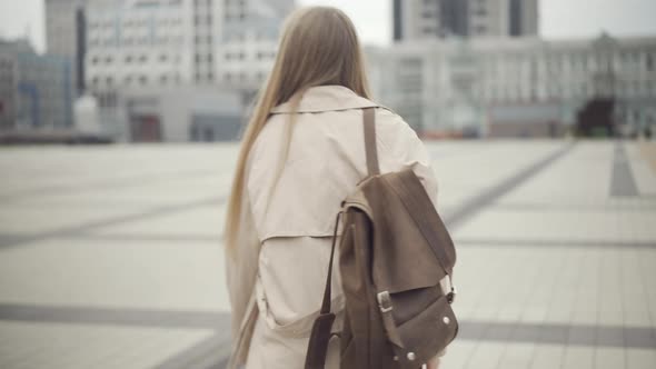 Camera Follows Young Caucasian Woman with Backpack Walking Along City Square and Looking Around