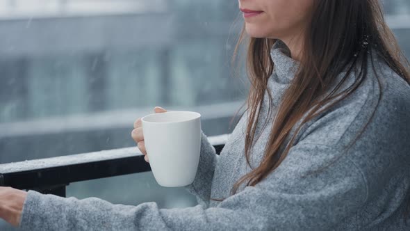 Caucasian Woman Stays on Balcony During Snowfall with Cup of Hot Coffee or Tea