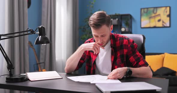 A Tired Young Man Sits at His Desk in His Room and Reviews His Apartment alt