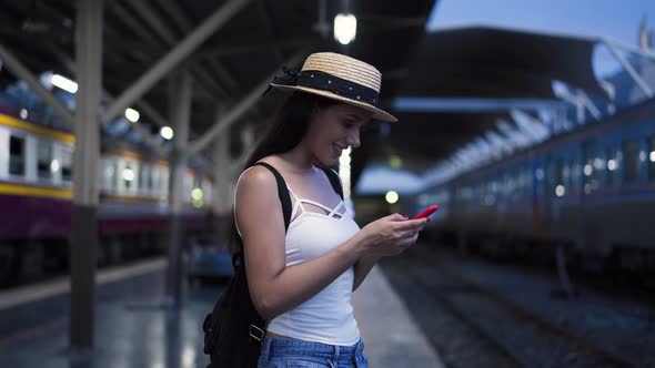 Young Brunette Woman Traveler Laughs While Playing a Mobile Phone in Train Station Platform