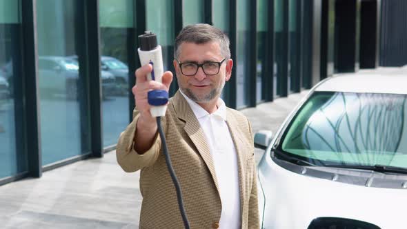 Older Man Stands with Charger Near His Electric Car