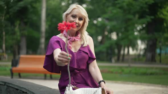 Medium Shot Positive Adult Woman Stretching Red Flower Looking at Camera Smiling Sitting at Fountain