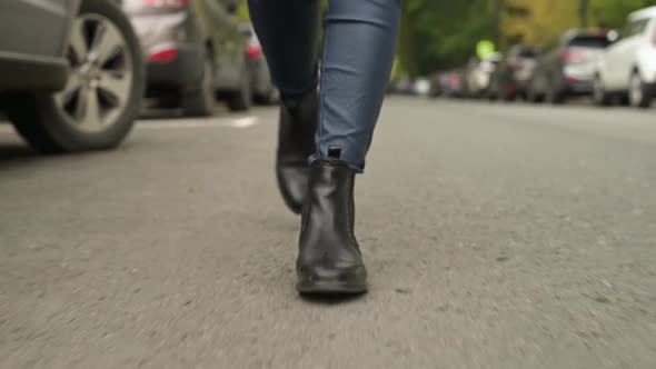 Female feet in leather black boots are walking on the asphalt, close-up