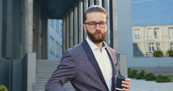 Businessman in Formal Suit Poses on Camera with Paper Cup with Coffee Near Modern Office Building