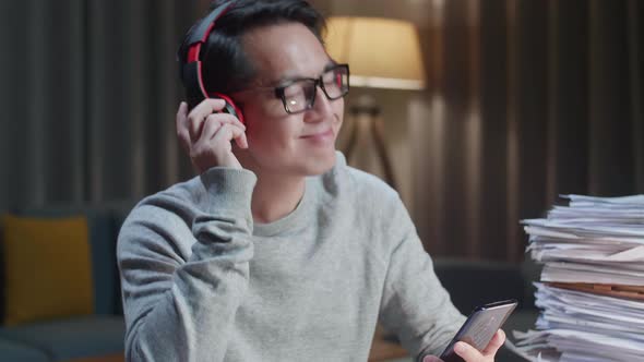Close Up Of Asian Man With Headphones Listening To Music On Smartphone After Working With Documents